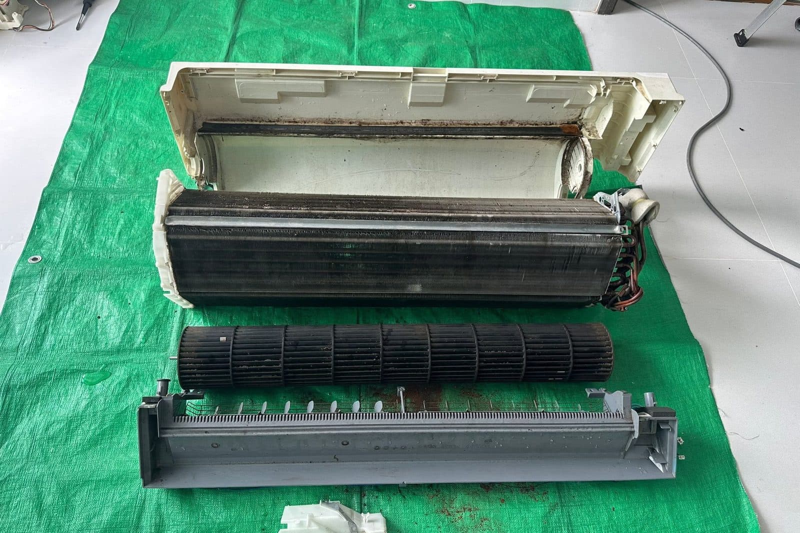 An aircon indoor unit stripped down on a work sheet — casing, evaporator coil, blower wheel and drainage tray, all still fouled